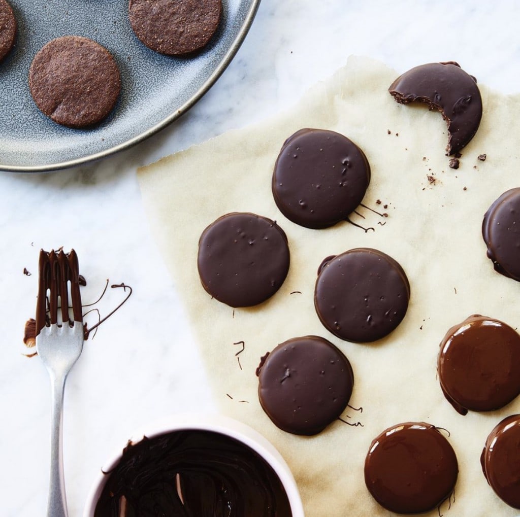 Chocolate covered cookies on parchment paper beside a bowl of melted chocolate, a fork that has been dipped in the chocolate, and a platter of cookies waiting to be dipped, all on a white counter.
