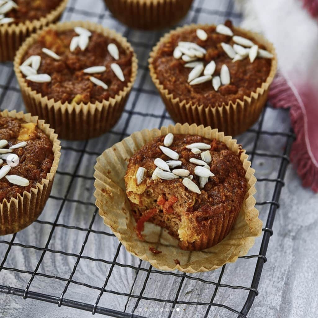 brown muffins topped with sunflower seeds in parchment paper cupcake wrappers cooling on a wire rack on a light gray wooden surface