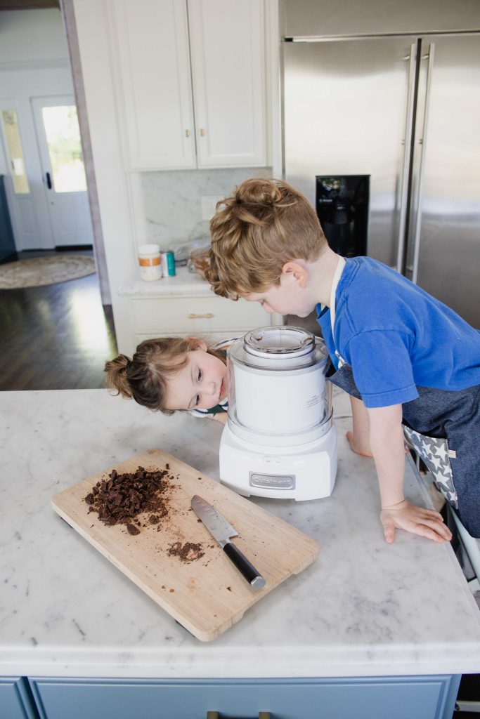 In a home kitchen, a young boy and young girl stare at a white ice cream machine. A wooden cutting board with chopped up cookies lays nearby on the counter.