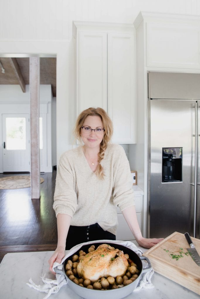 women smiling at camera stands in white home kitchen in front of a large pot containing a whole roasted chicken and potatoes