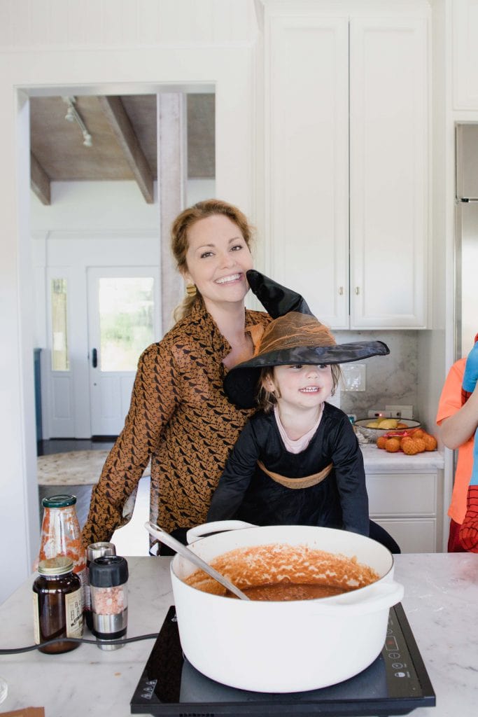 woman smiles at camera while standing in front of a counter that holds large white pot with chili contents. A small girl in a black and orange witch costume stands in front of her looking off camera.