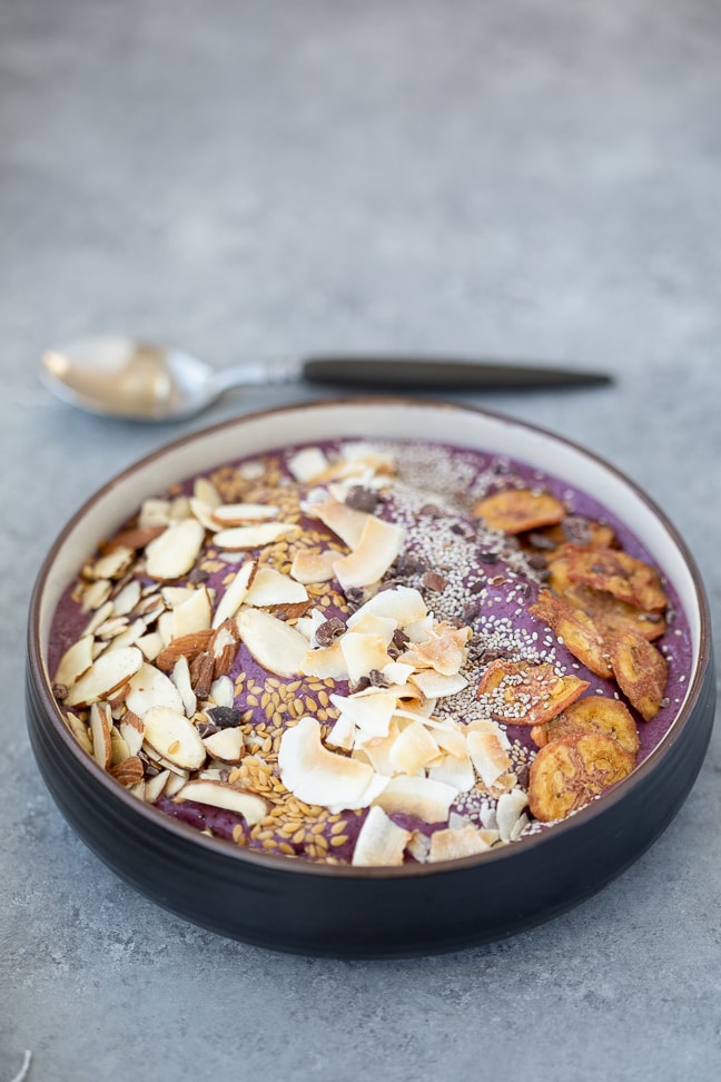 gluten-free acai bowl topped with a variety of grain free options such as almonds, coconut, chia seeds and plantain chips on a gray counter with a spoon in the background