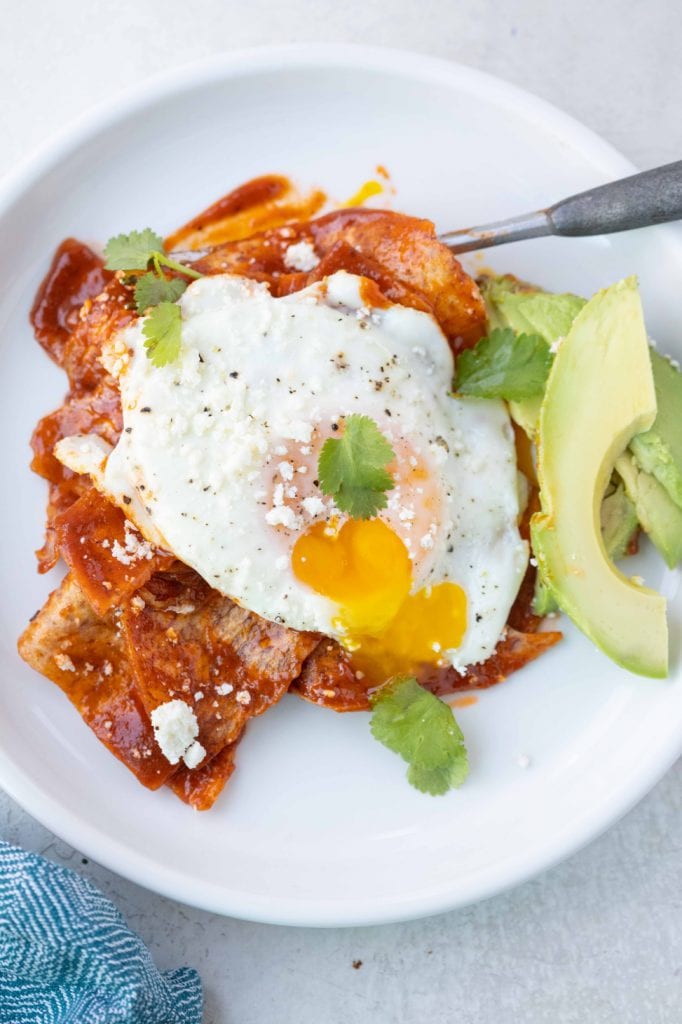 chips smothered in red sauce with an egg over easy on top with a side of avocado with cilantro as garnish, all on a white plate