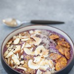 gluten-free acai bowl topped with a variety of grain free options such as almonds, coconut, chia seeds and plantain chips on a gray counter with a spoon in the background