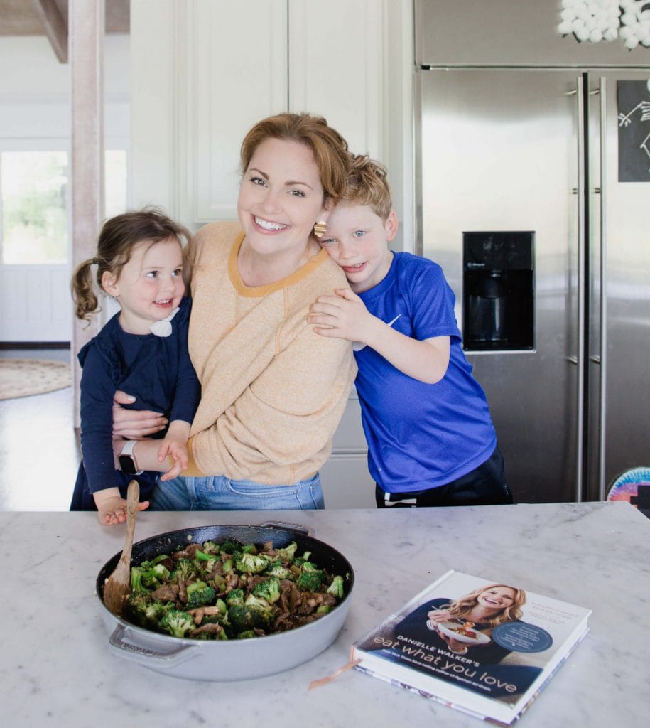 Woman stands in home kitchen embracing her daughter while she is embraced by her son. They are all smiling. On the counter rests a pot of beef and broccoli stir fry and her cookbook lays close by.