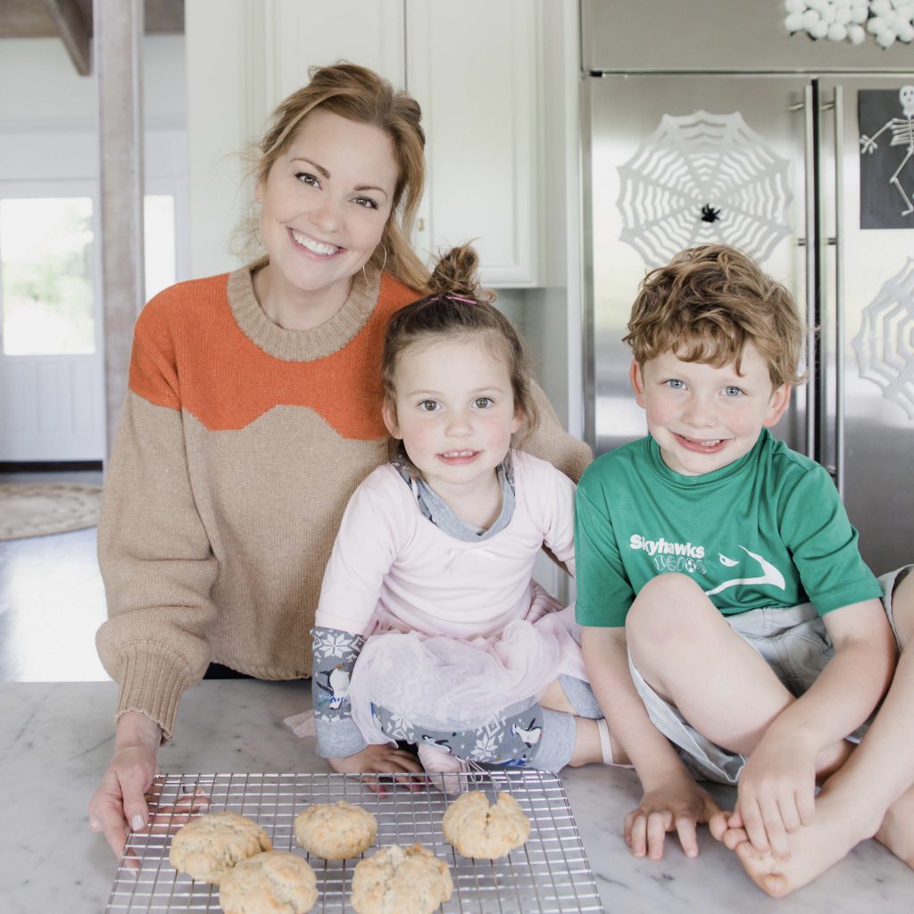 two children sit on kitchen counter by a wire rack with biscuits cooling. Mother stands behind them smiling at camera.