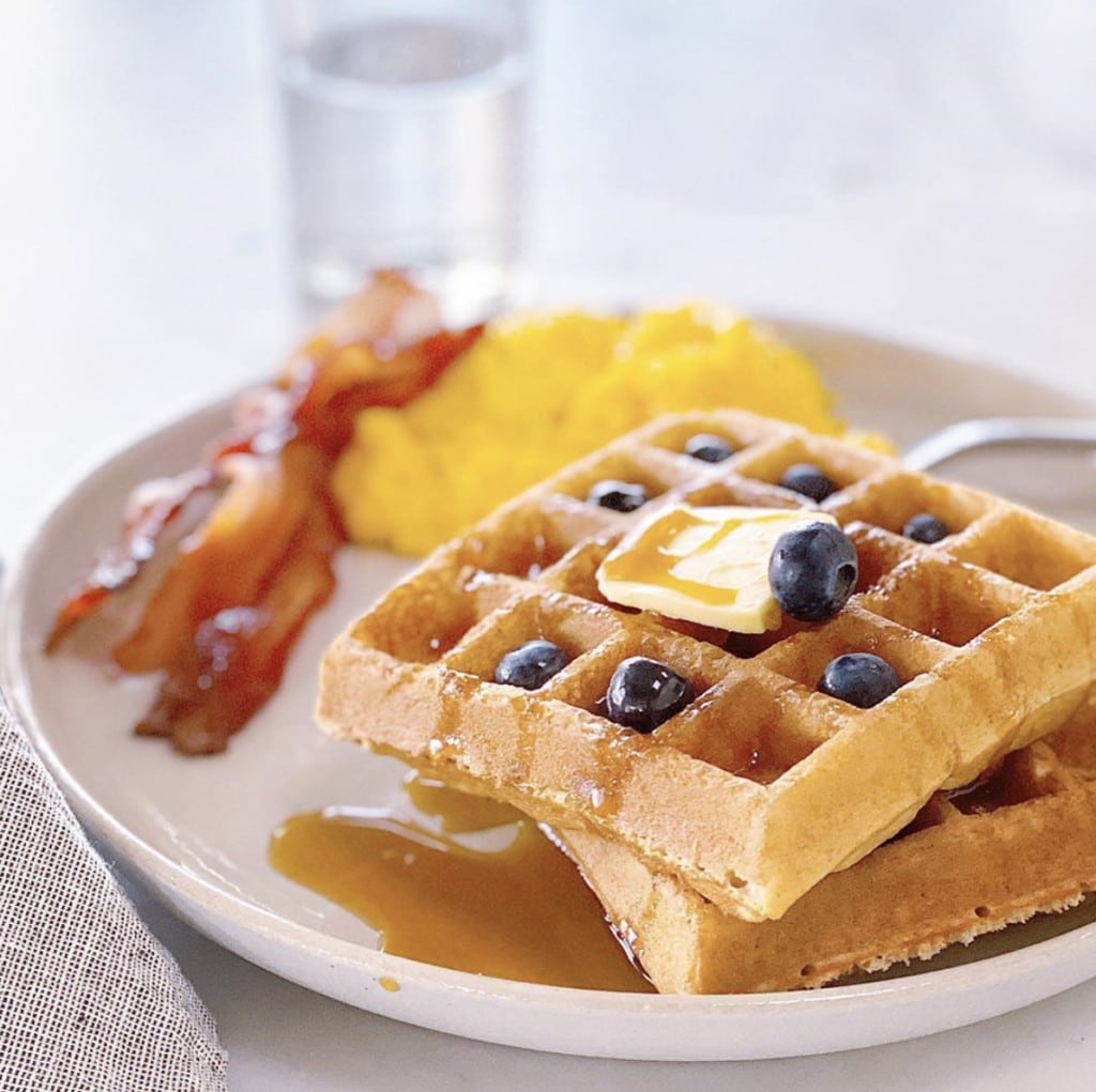 Waffles topped with syrup, blueberries and butter, eggs and bacon on white plate with a glass of water in the background, all on a white surface
