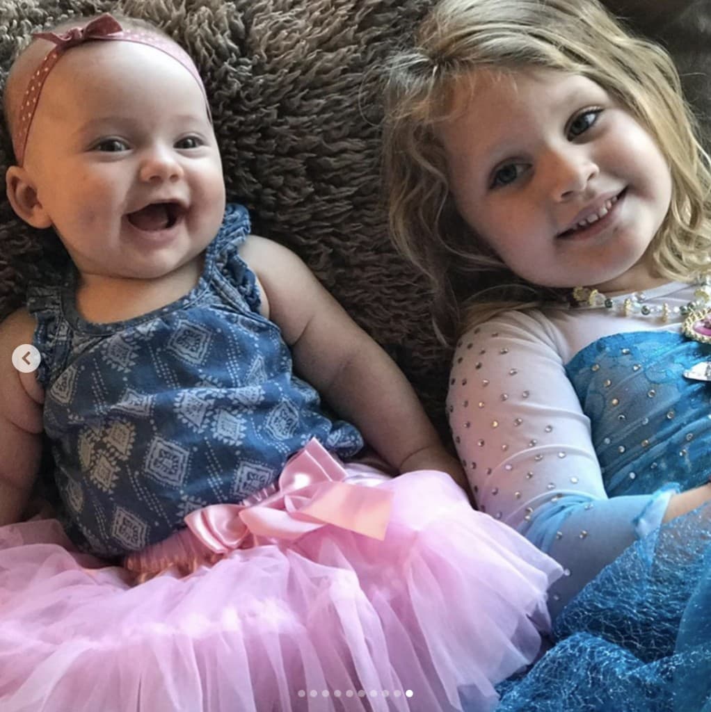 smiling blonde female toddler lays on a beige shaggy carpet next to a gleeful infant