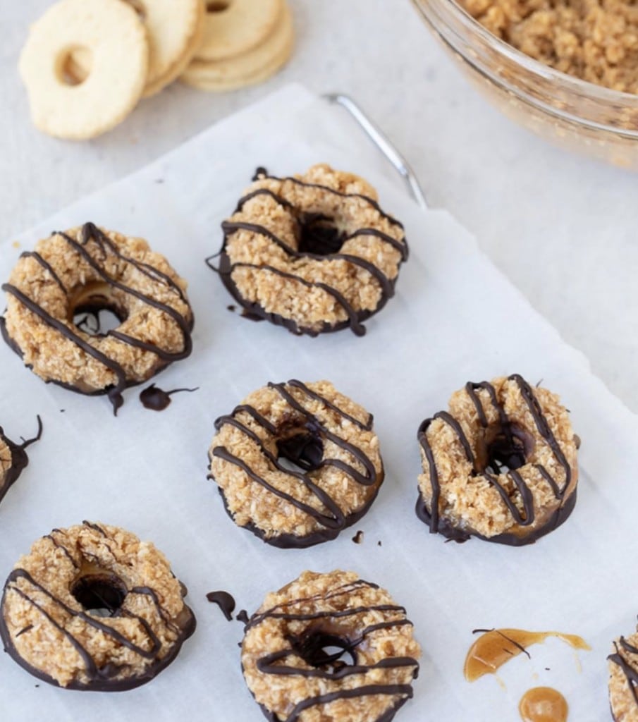 coconut caramel cookies drizzled with chocolate on a parchment paper. A glass bowl with coconut and caramel mixture and shortbread cookies in background - all on a white counter.