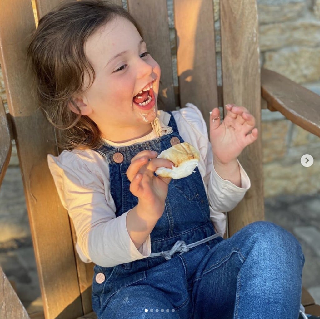 cheerful young girl sits in wooden chair outdoors enjoying a roasted marshmallow