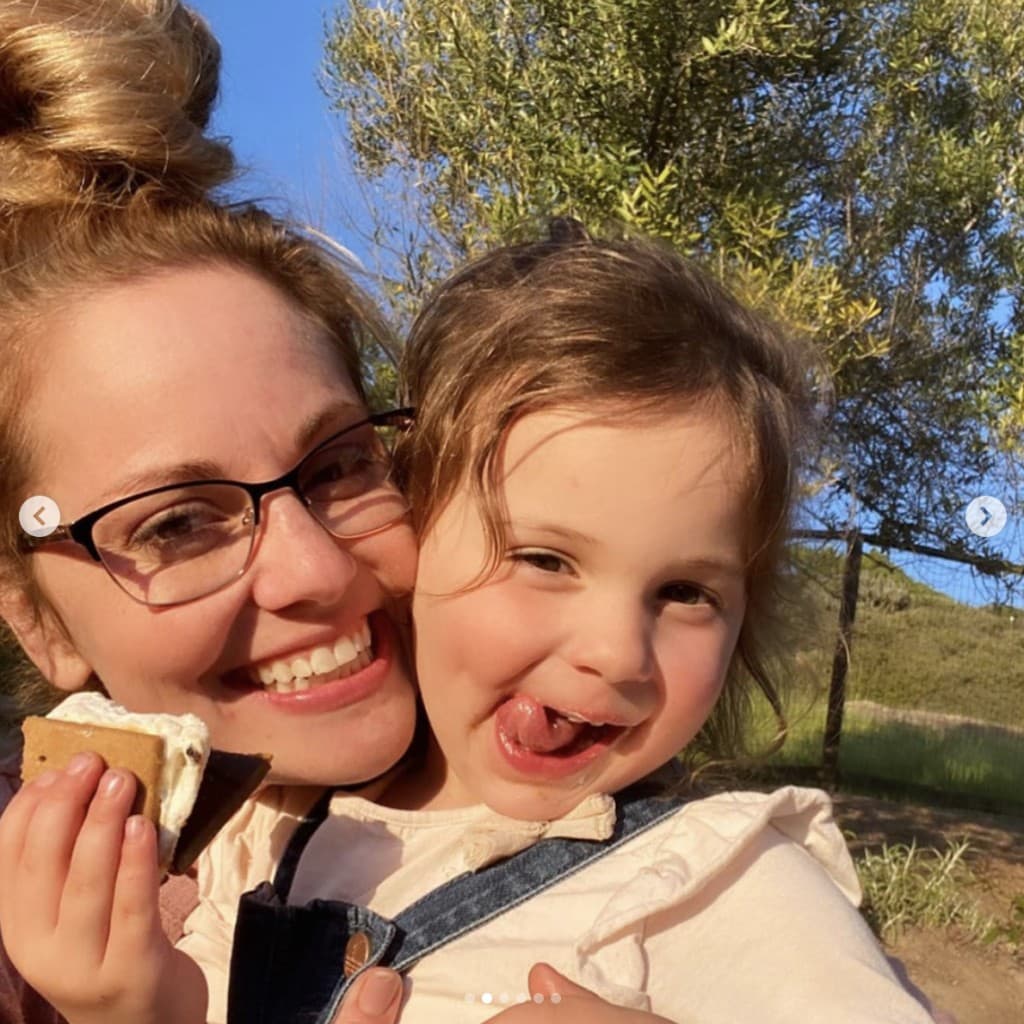 Cheek to cheek, a woman and young girl smile playfully into camera. Young girl holds a s'more in her hand.