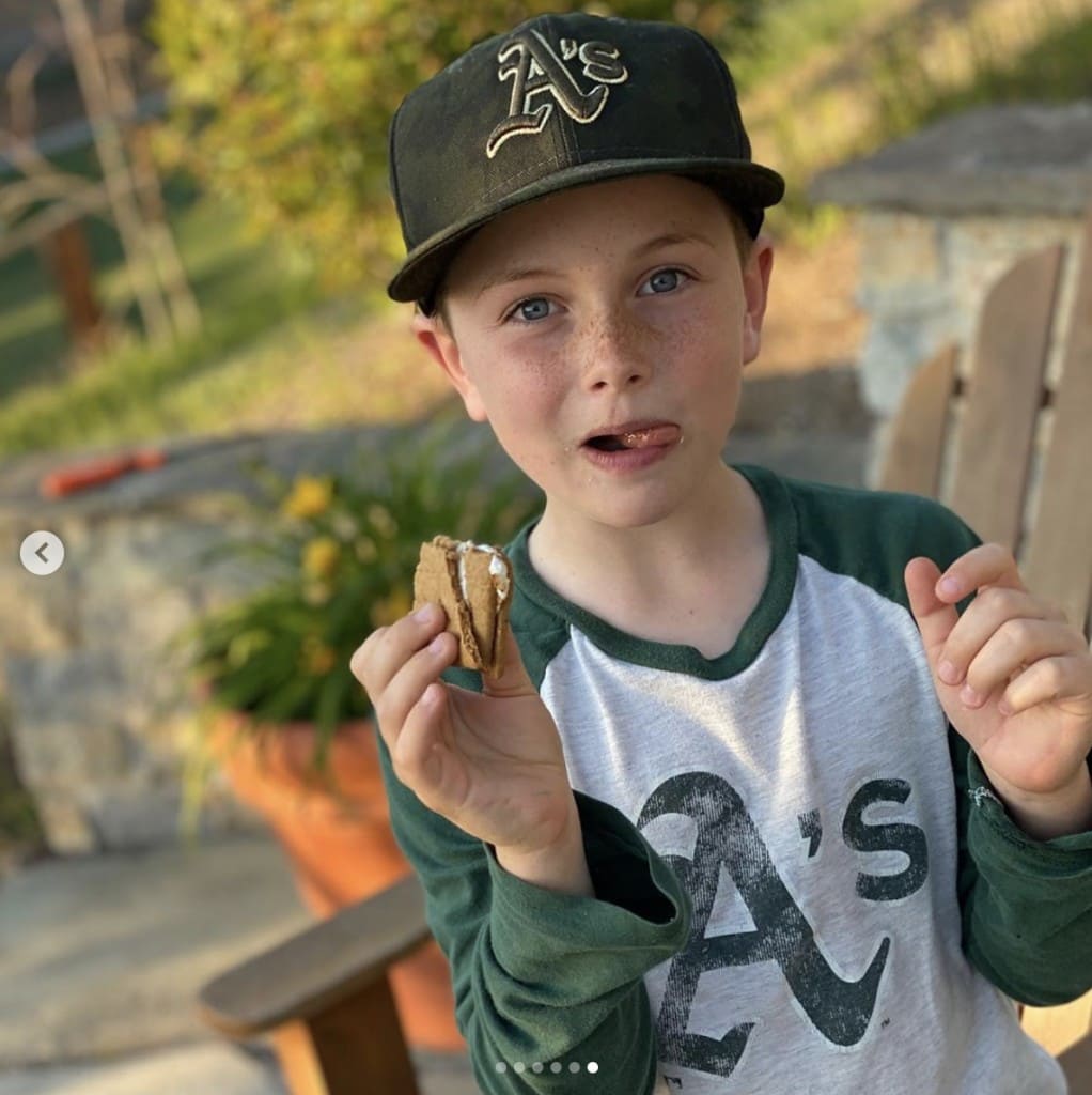 male child wearing baseball cap looks into camera while eating his s'more