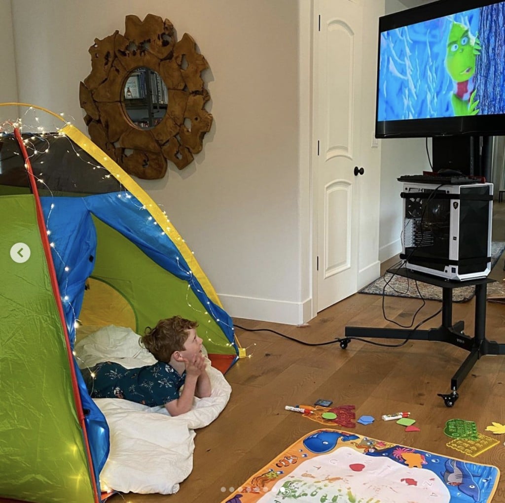 young boy lays in an indoor tent gazing up at a television stand; television is playing a version of The Grinch Who Stole Christmas.