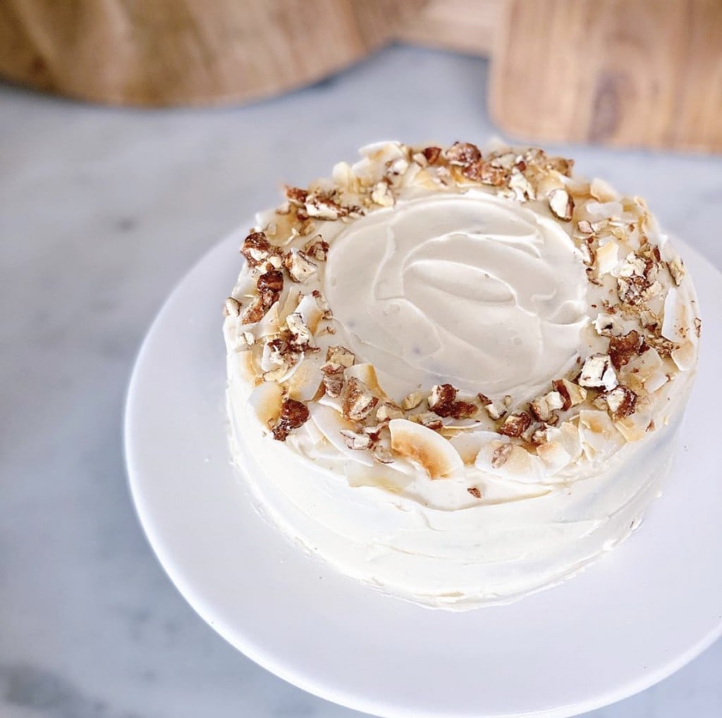 carrot cake with cream cheese decorated with a variety of nuts and coconut flakes. Cake is on a white platter on a white counter.