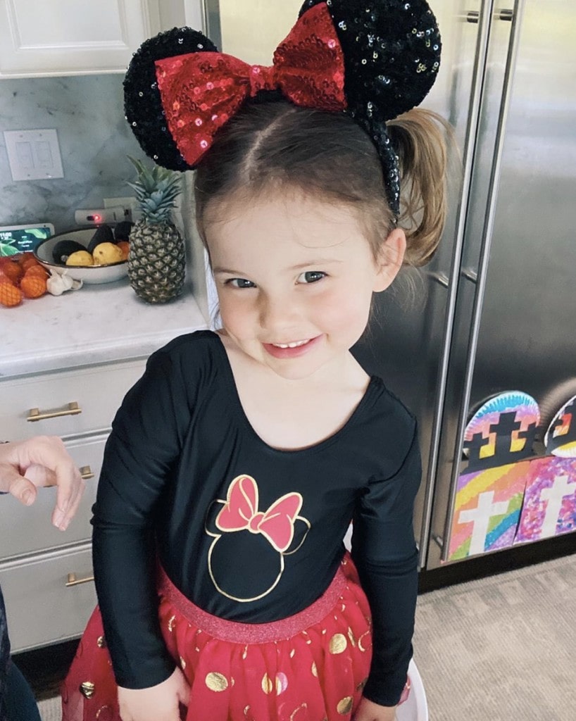 little girl stands in home kitchen and smiles at camera while wearing red and black Minnie Mouse outfit and sequin Minnie Mouse ears with a giant red bow