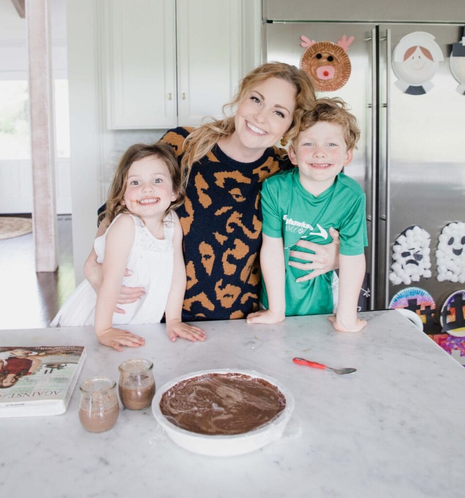 Woman embraced children in home kitchen. They are all smiling happily at camera while displayed a chocolate pudding pie.