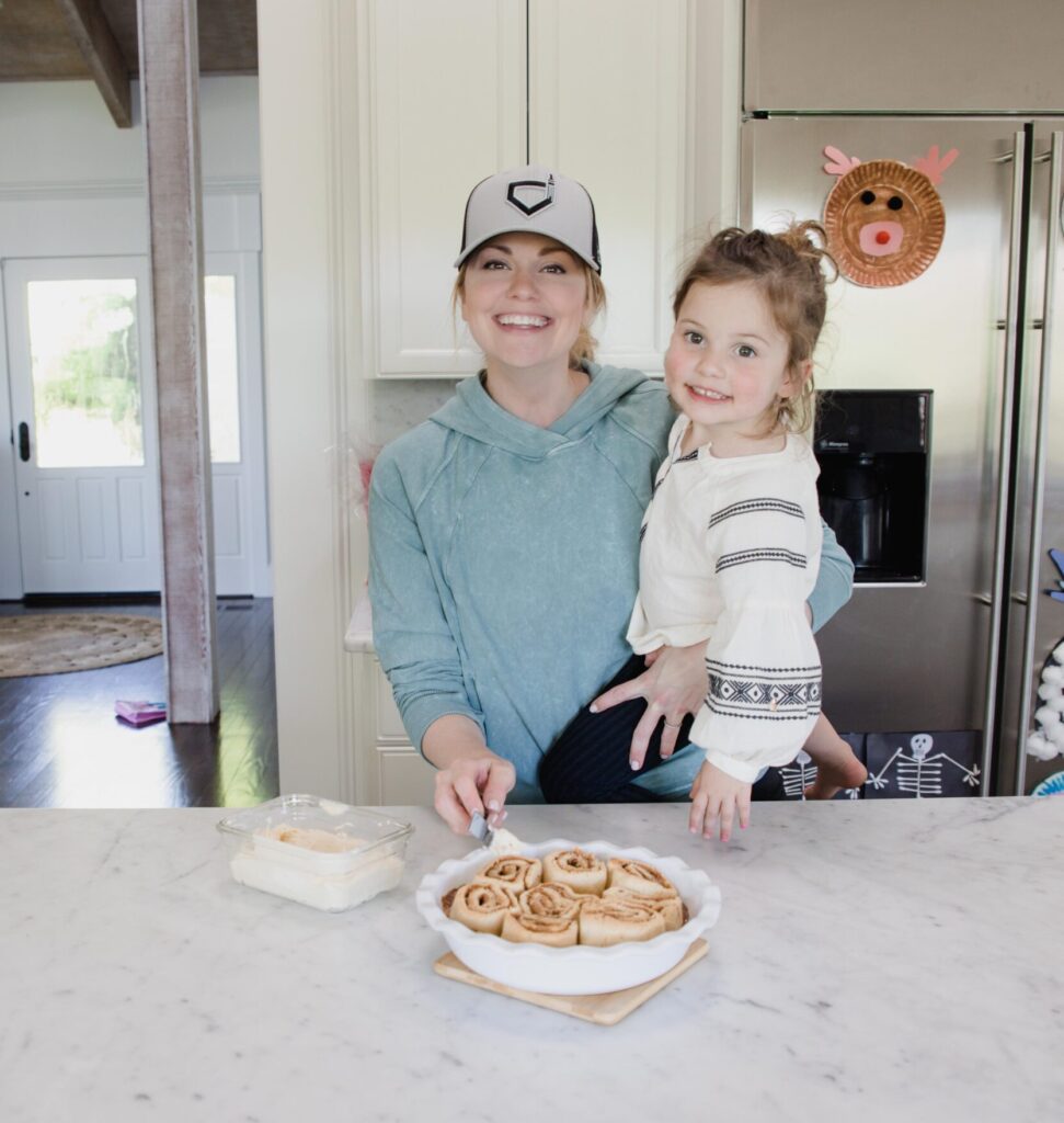 woman carries toddler daughter at hip while they both smile at the camera. Woman is preparing to ice freshly made cinnamon rolls with her free hand