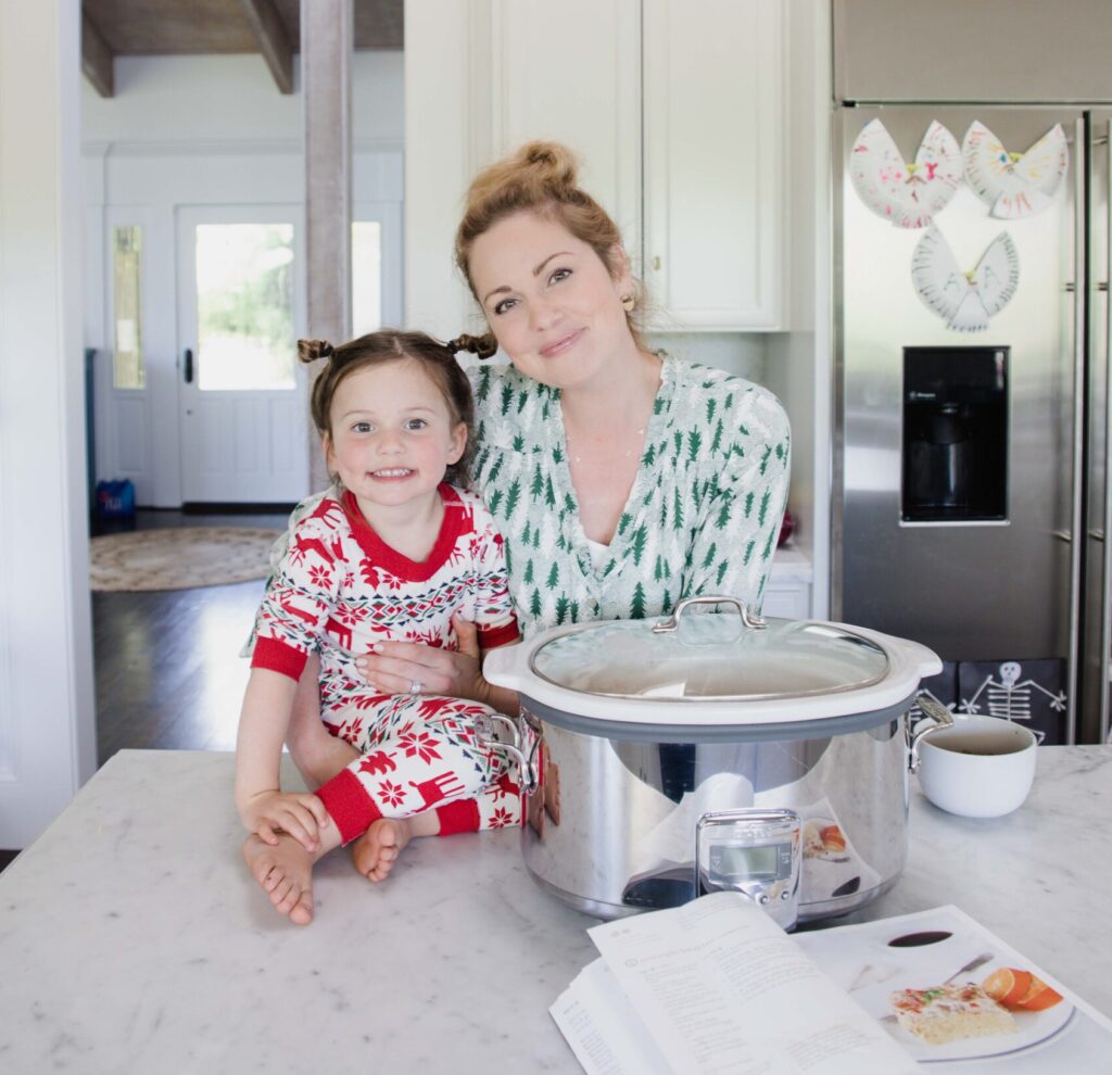 smiling woman stands in kitchen holding her young daughter, who sits on kitchen counter smiling at camera as well. A slow cooker sits on counter top in front of an open cookbook