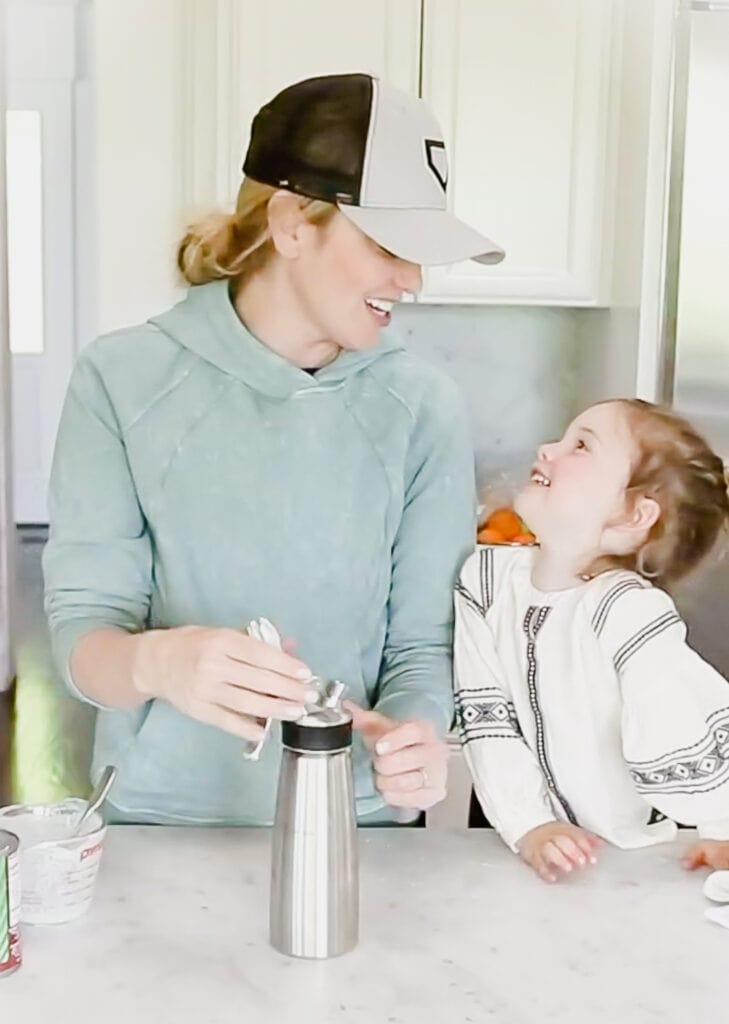 Woman and toddler daughter smile lovingly at each other while they stand in home kitchen preparing dairy free whipped cream
