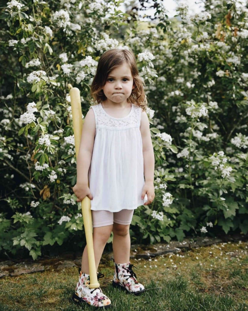 Young girl stands outdoors in front of a flowery bush, seriously staring in camera holding a toy baseball bat. She wears a white tank top, shorts, and flower patterned white combat boots