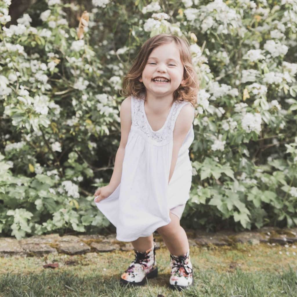 Young girl poses in front of flowery bush smiling gleefully at camera. She wears a white tank top, beige shorts and flower patterned white combat boots