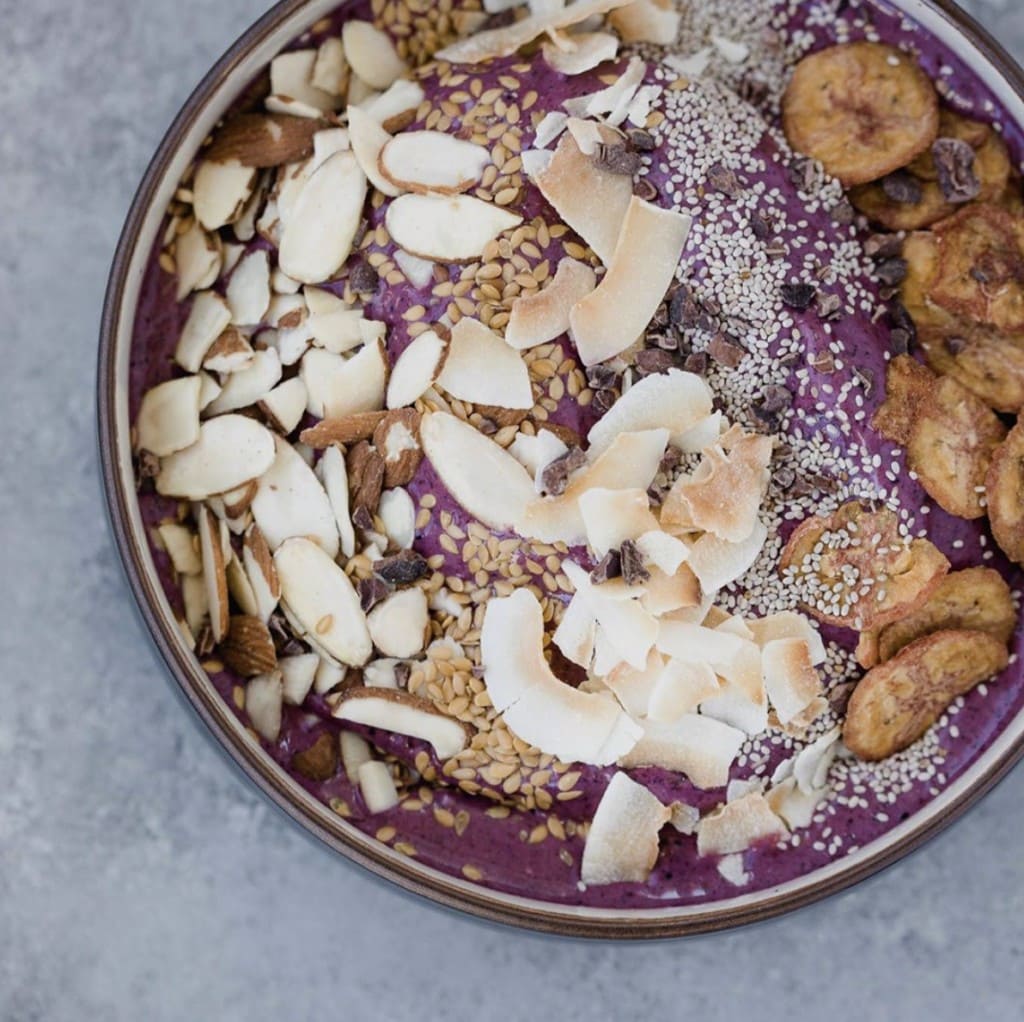 Purple acai smooth bowl topped with a variety of crunchy additions such as plantain chips, coconut flakes, slivered almonds, chia seeds, etc.