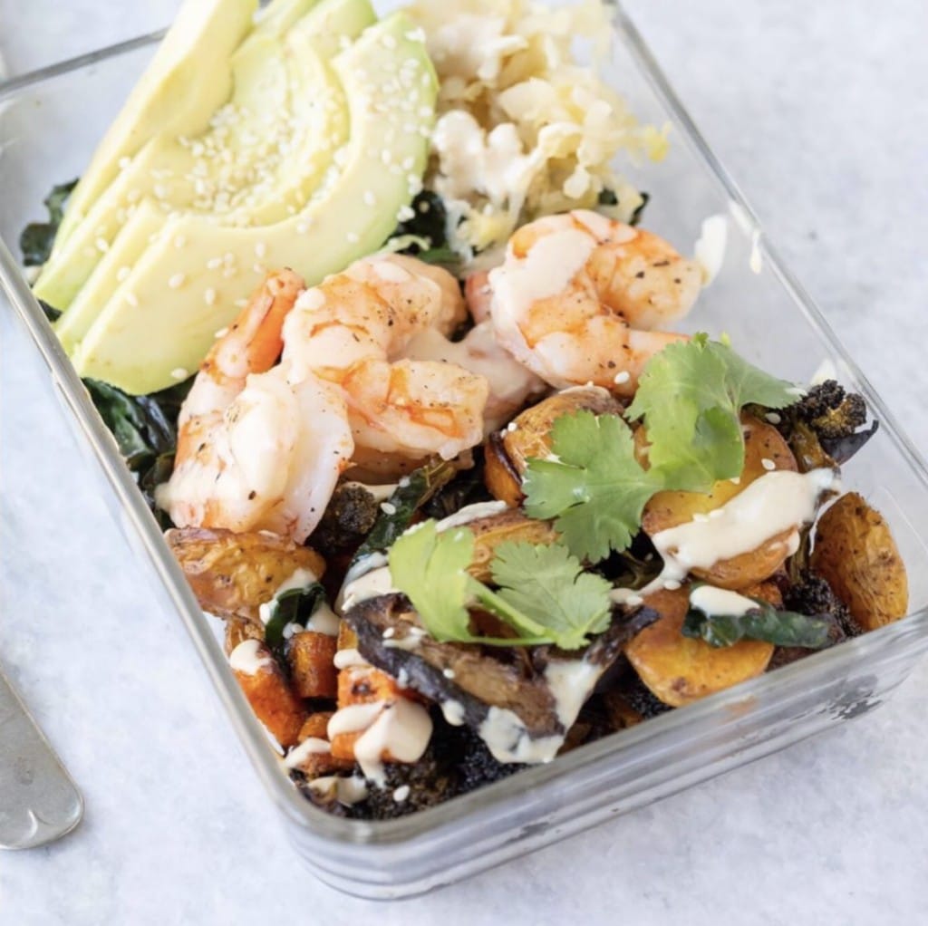 Up close view of roasted vegetables in a glass bowl - topped with fresh avocados, shrimp, fresh cilantro and a drizzle of tahini dressing