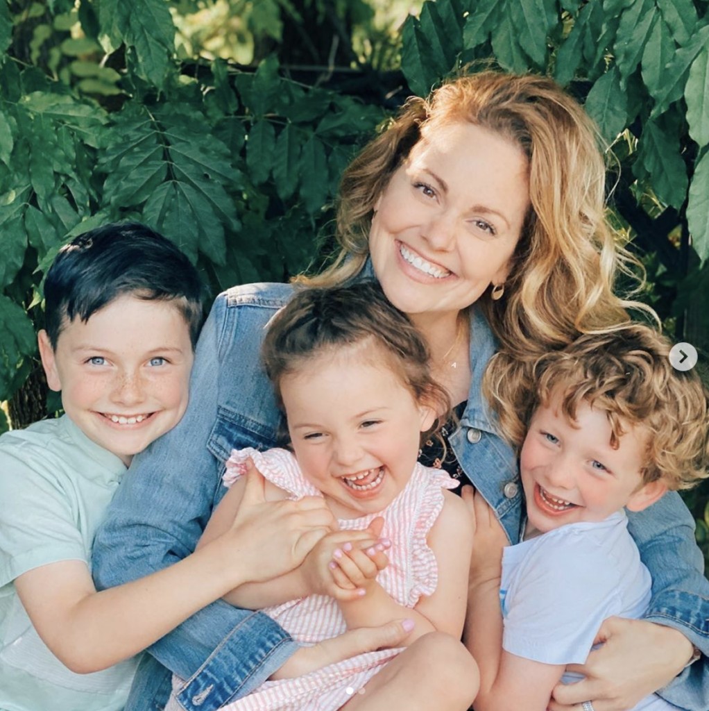 Woman smiles joyfully at camera while embracing three gleeful children