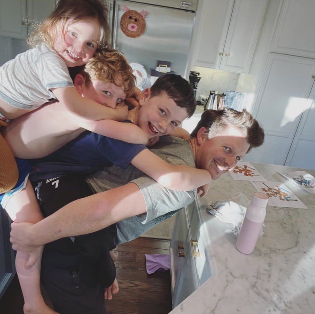 Father stands in home kitchen smiling at camera with three children playfully stacked on his back