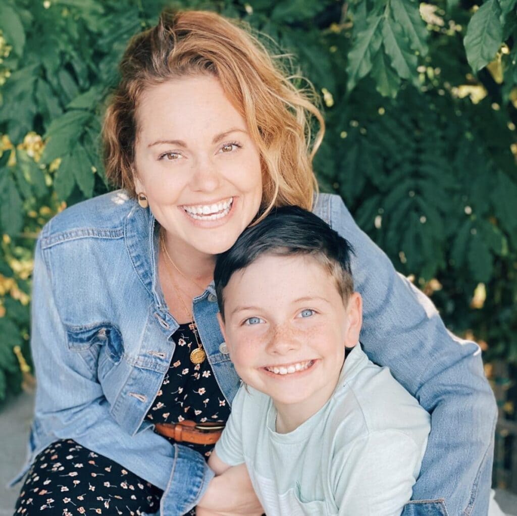 woman wearing a jean jacket and summer dress embraces son while they both smile directly at the cam