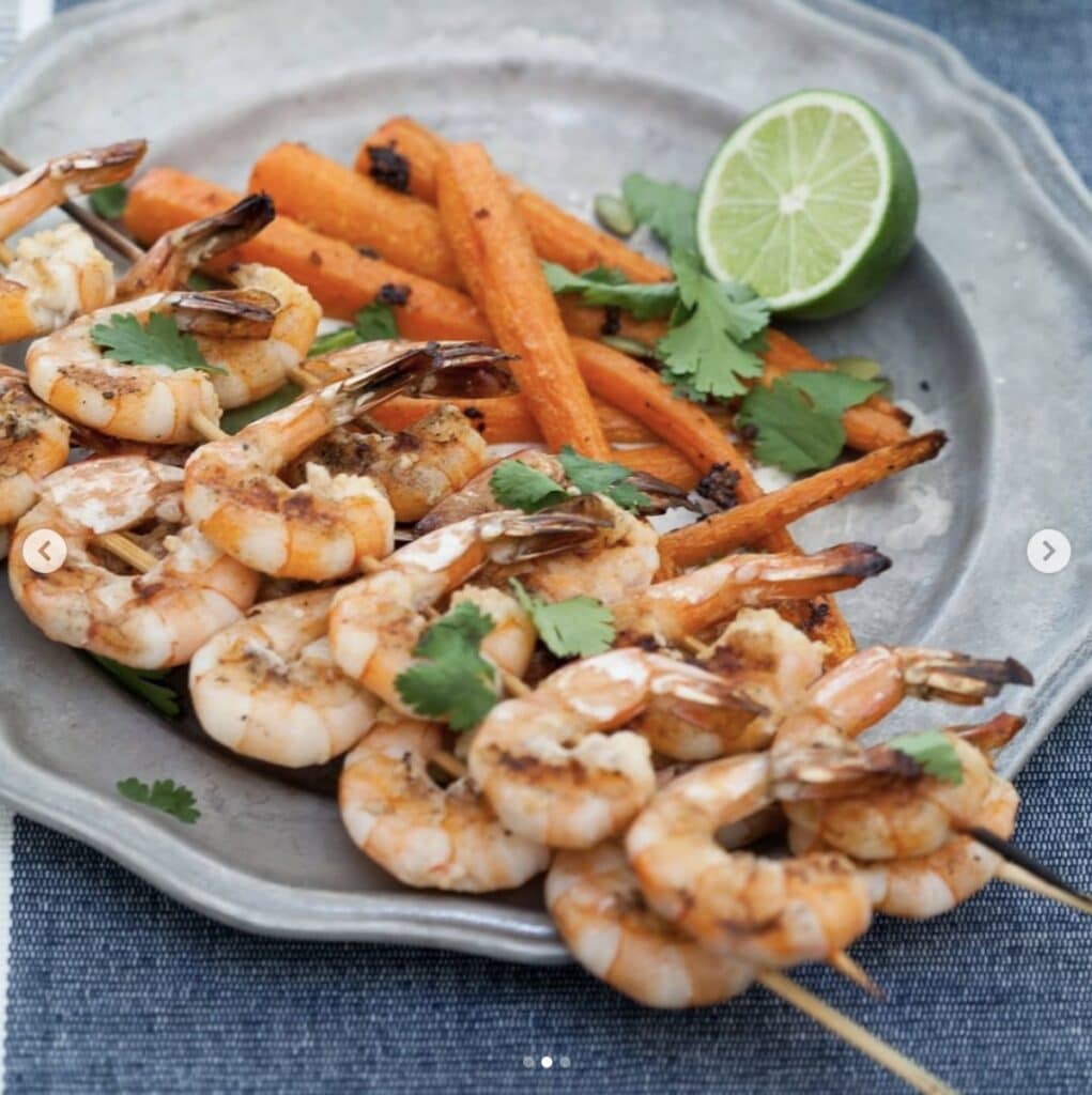 Shrimp on wooden skewers with a side of carrots, garnished with cilantro and lime on a gray dish