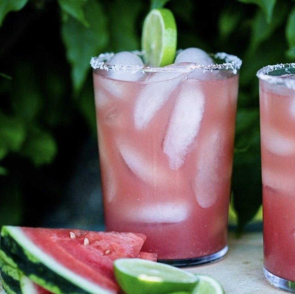 glass of watermelon margarita filled with ice, a salted rim and a slice of lime as a garnish next to watermelon and lime slices on a white table outdoors