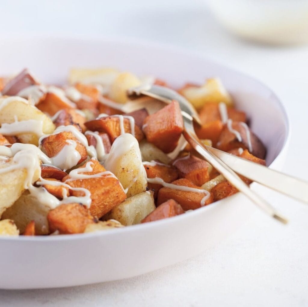roasted pineapple and sweet potatoes drizzled with cashew cream in a white bowl with gold colored spoons on a white table