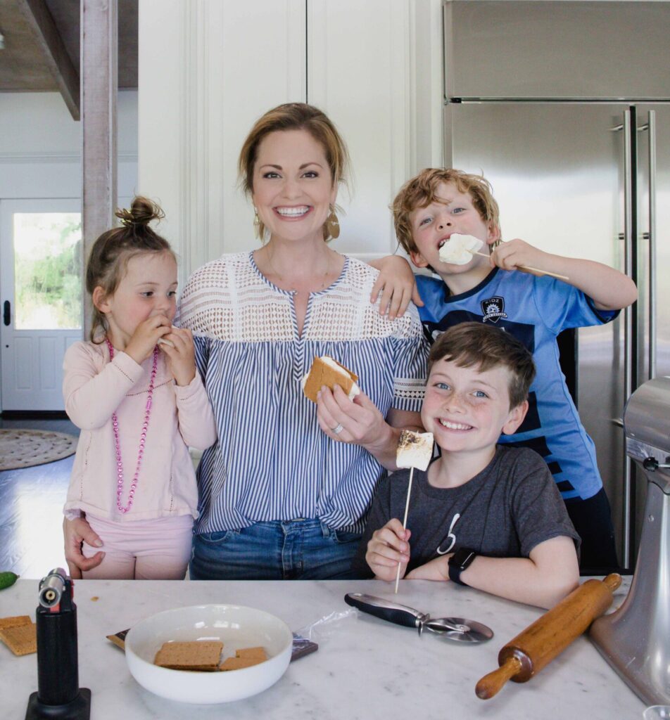 Woman stands in home kitchen smiling at camera while she and her three kids enjoy homemade s'mores with her children.